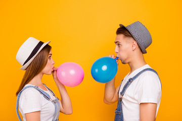 Young happy couple in love wearing hats blowing air balloons looking confused of yellow background,...