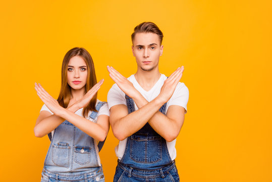 Portrait of lovely young beautiful, confident students couple standing together seriously showing stop no sign with their arms, isolated over yellow background