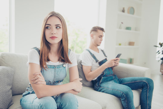 Young attractive beautiful cute students couple wearing overall sitting home on sofa, sad girl being ignored by guy who is chatting in his smartphone