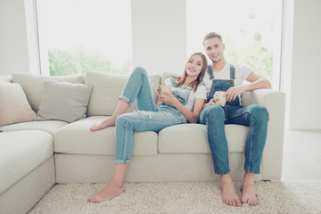 Obraz premium Young attractive cute couple wearing overall sitting home on sofa bearfoot, drinking coffee