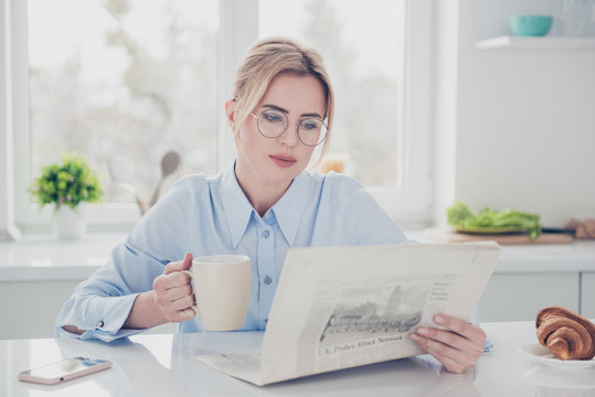 Adult adorable woman office executive worker wearing eye-glasses keeping in hands and reading newspaper early in the morning having a drink with croissant in light kitchen