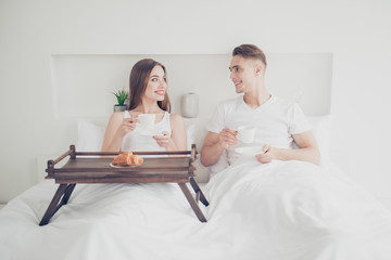 Portrait of young attractive cute couple sitting together in bed, covered with blanket, smiling, having breakfast, drinking coffee, eating croissants