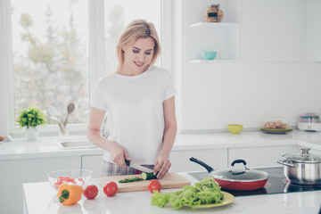 Obraz na płótnie Canvas Adorable attractive adult smiling woman with manicure chopping cucumber on board with knife, cooking nice salad wearing pajama. Chopped pepper, tomatoes, yellow pepper lying on white table