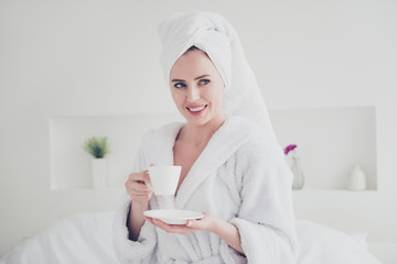 Young caucasian cute smiling woman wearing white bathrobe and turban drinking from cup holding plate in hand in bedroom with white interior. Flowers in vases bed sheets on background