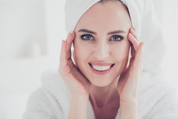 Close up portrait of young cute adult woman looking straight at camera smiling with her teeth wearing white bathrobe and turban keeping hands and fingers near eyes wrinkles in white interior