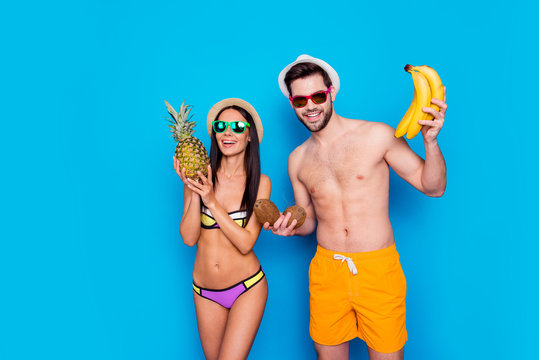 Young and charming lady dressed in swimsuit holds a pineapple standing beside a man in yellow shorts with tropical fruits on his hands isolated on blue background. Vitamins and healthy summer concept