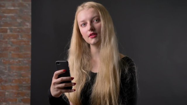 Young cute blond girl with pink lipstick is typing message on smartphone, communication concept, gery background