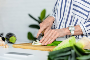 cropped image of grey hair woman cutting fresh green onion on wooden board in kitchen
