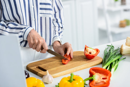 cropped image of grey hair woman cutting red bell pepper on cutting board in kitchen