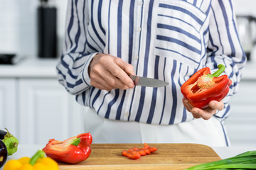 cropped image of grey hair woman cutting red bell pepper on wooden board in kitchen