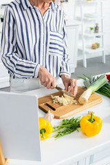 cropped image of senior woman cutting uncooked leek on wooden board in kitchen
