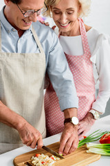 smiling senior wife hugging husband while he cutting green onion at kitchen