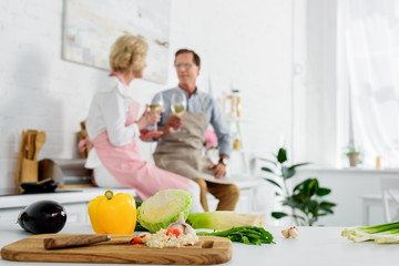 close-up view of fresh vegetables in wooden cutting board and senior couple drinking wine behind in kitchen