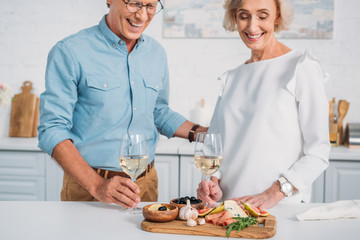 cropped shot of smiling senior couple holding glasses of wine and looking at delicious snacks at home