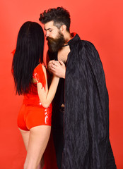 Vampire in cloak and sexy devil girl holds hands. Couple in love, perfect match. Couple on pensive faces play role game. Man and woman dressed like vampire, demon, red background. Halloween concept.
