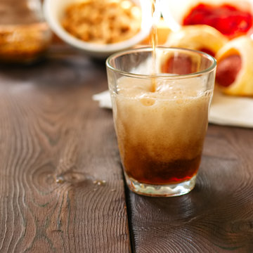 Pouring dark beer into glass with pigs in a blanket, ketchup, nuts and mustard on background. Square image.