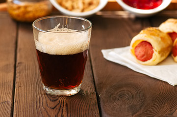 Close up of dark beer in a glass with pigs in a blanket, ketchup, nuts and mustard on background