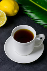 A cup of tea, a lemon and a green leaf on a dark table