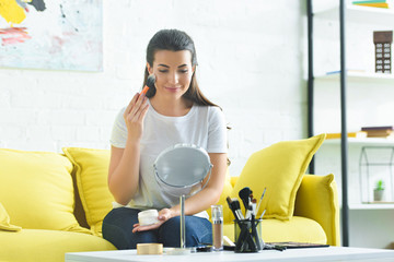 portrait of smiling woman applying face powder while doing makeup at home
