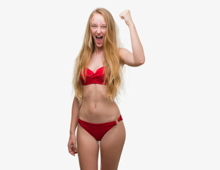 Obraz premium Blonde teenager woman wearing red bikini angry and mad raising fist frustrated and furious while shouting with anger. Rage and aggressive concept.