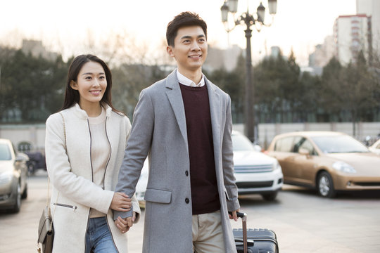 Cheerful young Chinese couple holding hands walking with wheeled luggage