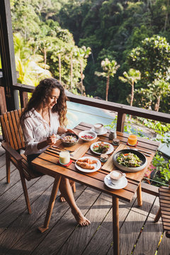Woman enjoying breakfast on terrace with amazing jungle view. White shirt and jeans short