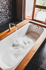 Fototapeta na wymiar Water filling bath with foam. Modern style of bathroom with wooden edge and black walls