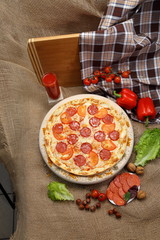 Pizza thin with smoked sausage, cheese, spices and tomatoes on a wooden stand with tomato juice in a glass sink. Cherry tomatoes and sweet peppers, lettuce leaves. Close-up