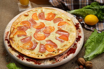 Pizza thin with sausage, bacon, spices and tomatoes on a wooden stand with orange juice in a glass sink. Cherry tomatoes and sweet peppers, lettuce leaves. Close-up