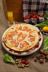 Pizza thin with sausage, bacon, spices and tomatoes on a wooden stand with orange juice in a glass sink. Cherry tomatoes and sweet peppers, lettuce leaves. Close-up