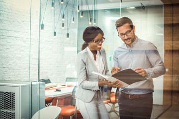 Through glass view of two smiling multi-ethnic business colleagues reviewing paper documents while standing in modern office
