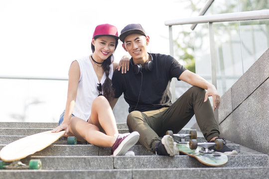 Portrait of cheerful young Chinese couple with skateboard sitting on stairs