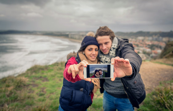 Portrait of young beautiful couple taking selfie photo in a smartphone with sea and dark cloudy sky on the background. Selective focus on phone.