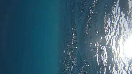 VERTICAL UNDERWATER, SLOW MOTION, LENS FLARE: Bright summer sunbeams peer through the tranquil ocean surface. Small gentle waves roll past the camera submerged in the crystal clear Pacific Ocean.