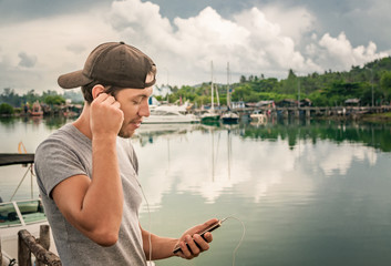 Young man walking near beautiful bay using phone and headphones for communicate with friends and...