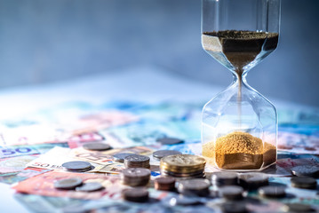 Sand running through the shape of hourglass on table with banknotes and coins of international...