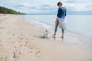 Woman walking with her dog breed Jack Russell Terrier on the sandy beach. No shoes on the water on...