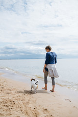 Woman walking with her dog breed Jack Russell Terrier on the sandy beach. No shoes on the water on a Sunny summer day. Rear view.
