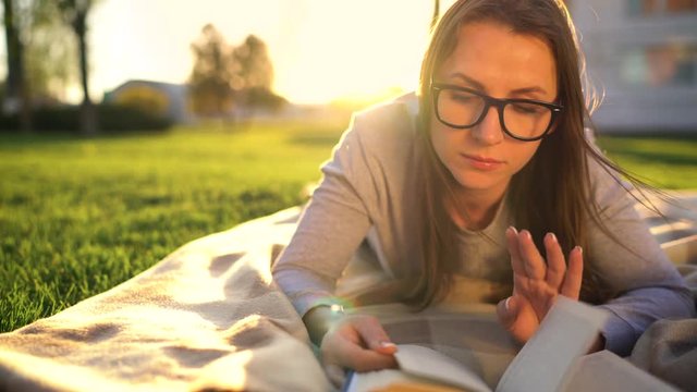 Girl in glasses reading book lying down on a blanket in the park at sunset