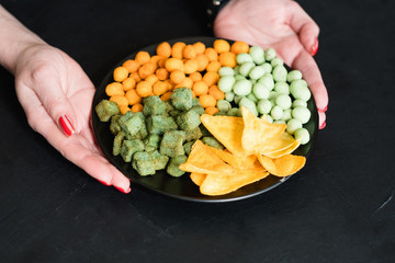 junk food and snacks concept. woman hands offering mix of salty spicy chips flavored peanuts and croutons on a plate.