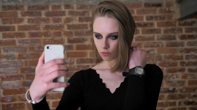 Young charming girl with smoky eyes make seifie on her smartphone, communication concept, brick background