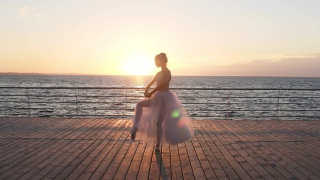 Beautiful silhouette of ballerina in long ballet tutu and pointe on embankment above ocean or sea at sunrise. Dancing, practicing with the sun rising on the background