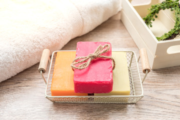 Three colorful soaps are placed in a basket, placed next to a towel on a wooden floor, and have a wooden box in the back.