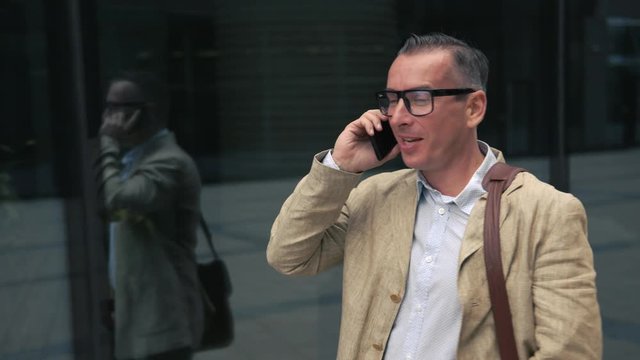 Man in casual wear and stylish glasses walking near modern building and talking on the phone