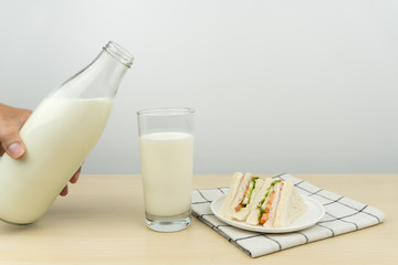 hand holding a milk bottle, a glass of milk and the club sandwiches in a white round dish on the wooden table.