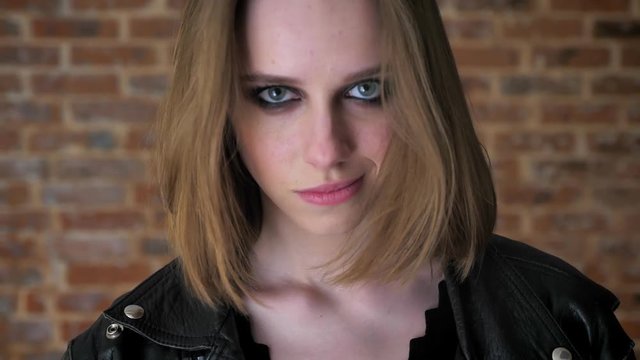Young sad girl with smoky eyes is watching at camera, smiling, biting lip, thinking concept, brick background