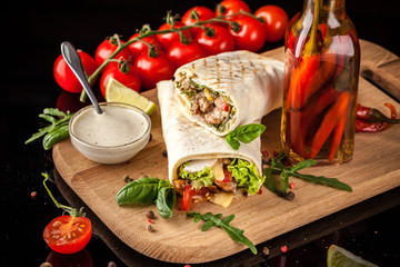 Mexican cuisine, burrito with chicken, cherry tomatoes, lettuce, mushrooms, rucola and chili peppers, on a black background. With salsa sauce. Copy space, selective focus