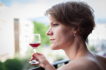 Young beautiful woman with a glass of red wine in hand, on the terrace of the house enjoys the summer sun
