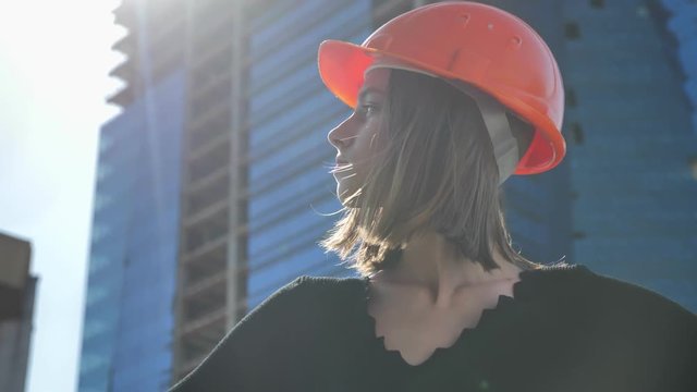 Young strong girl builder with hemlet is standing with hands on hips in daytime in summer, turning head, building concept, urban concept, building on background