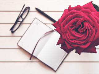 An open notepad with blank pages, black-rimmed glasses, a black pen and a large red rose are on a white wooden background.